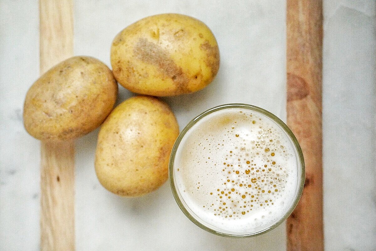 glass of potato juice sitting on counter with three yellow potatoes sitting next to it.