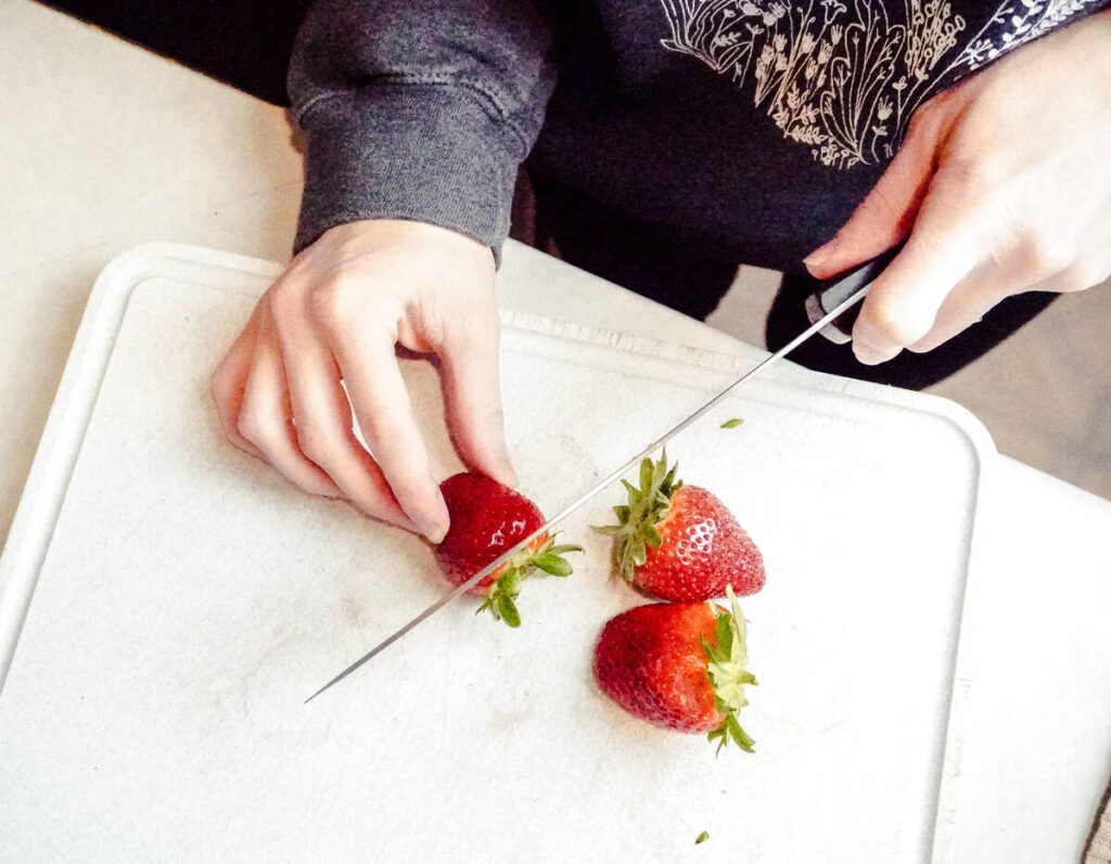 woman using a chefs knife on a gray cutting board to cut the top off of a strawberry