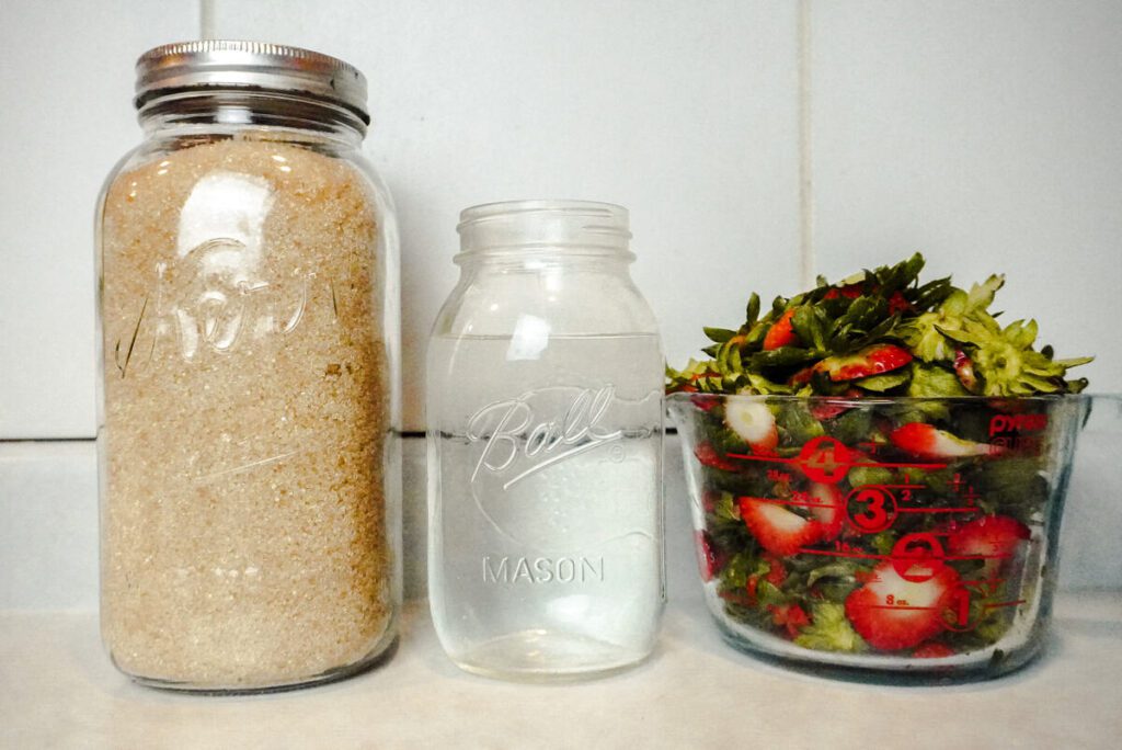 1//2 gallon jar organic cane sugar on counter top next to quart jar of water and liquid measure full of cut strawberry tops. 