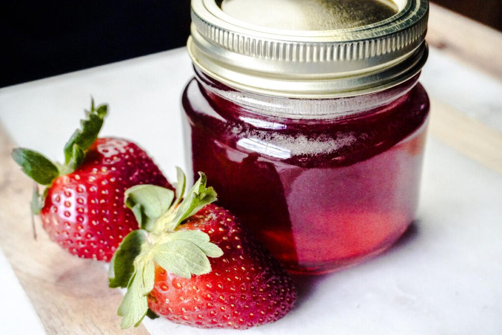 jar of strawberry top simple syrup on marble counter with 2 fresh strawberries next to the 1/2 pint jar. 