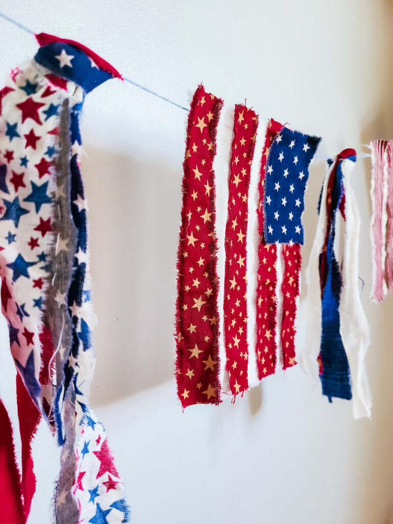 Close up photo of bunting flag banner hanging on a white wall. zoomed in on a red stripe flag with small gold stars and a blue small rectangle corner with white stars.