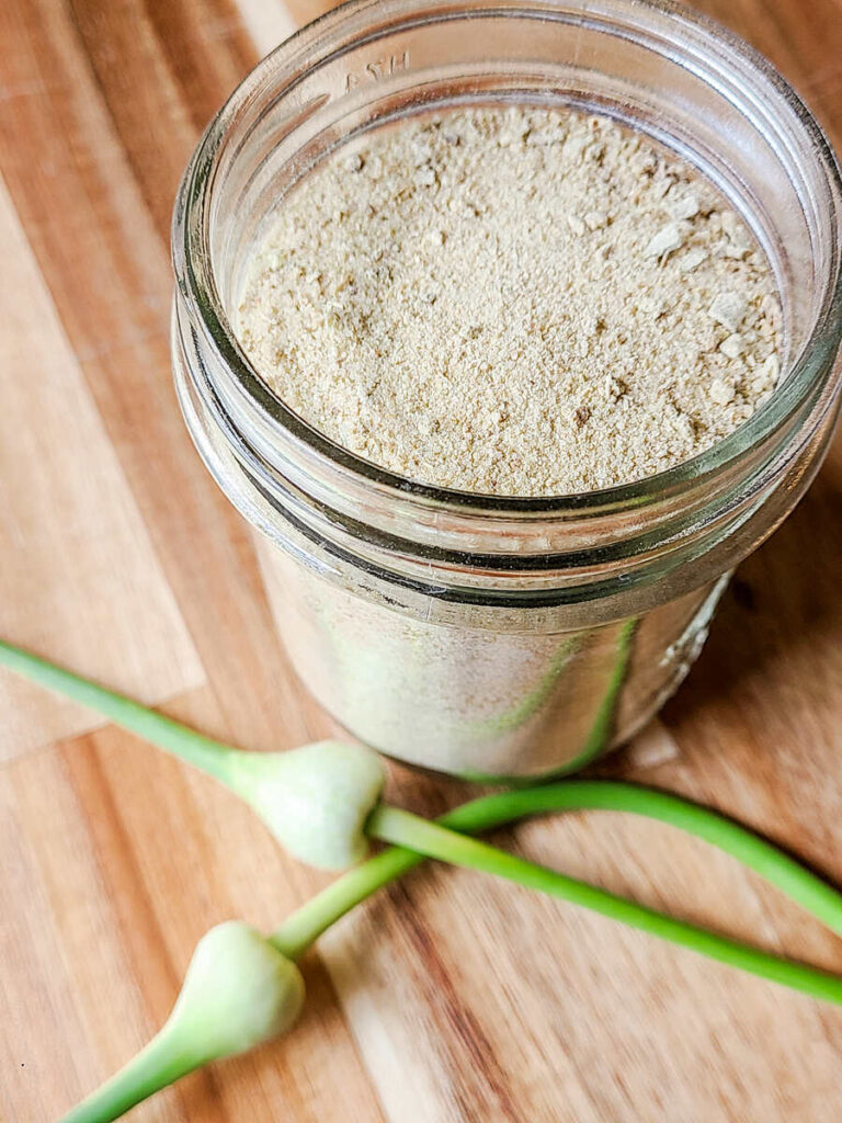 butcher block background, half pint jar full of brownish garlic scape salt. 2 garlic scapes sit next to the jar.