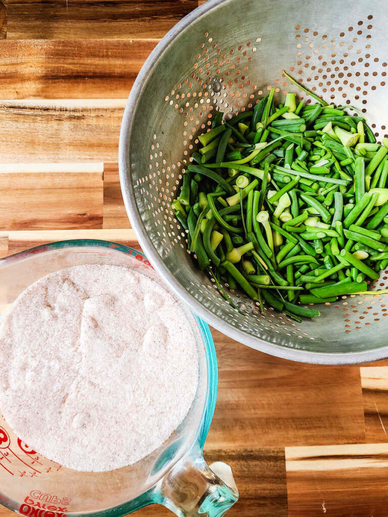 cut garlic scapes in gray colander next to bowl of salt on butcher block counter. 
