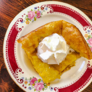 top view of square of German pancake topped with syrup and whipped cream on a red floral plate with a dark wood table back drop.