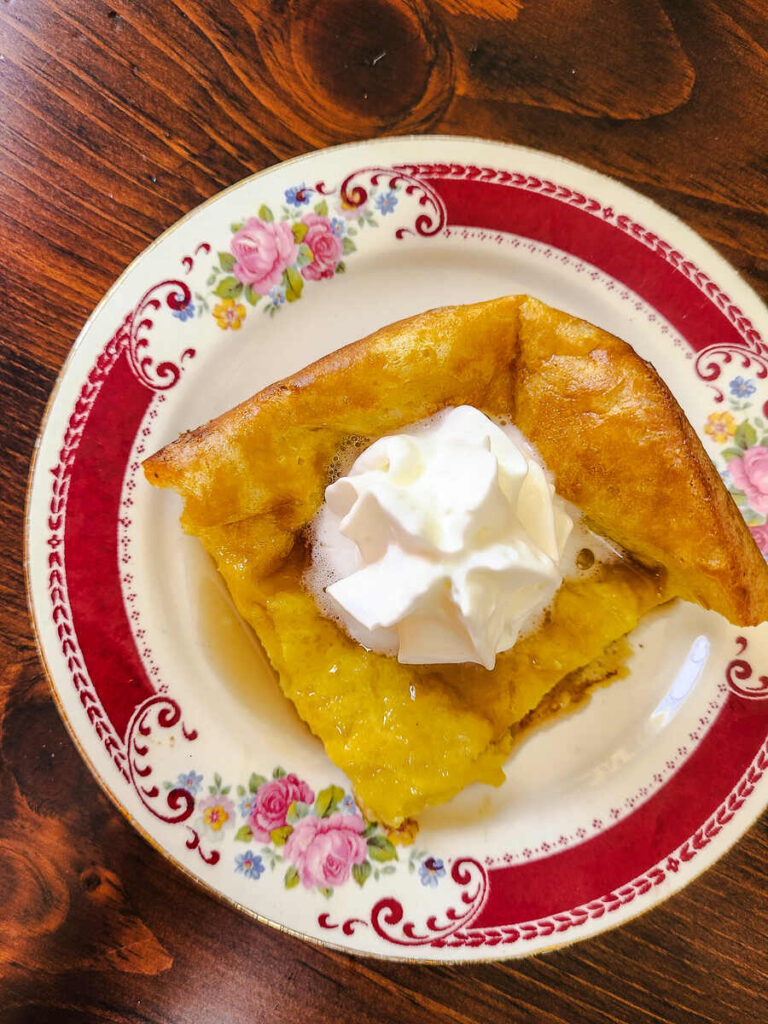 top view of square of German pancake topped with syrup and whipped cream on a red floral plate with a dark wood table back drop.