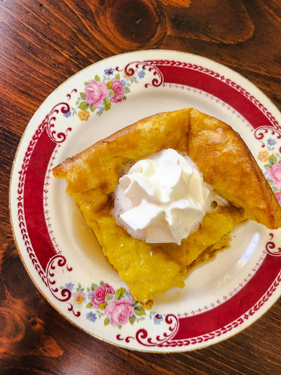 top view of square of German pancake topped with syrup and whipped cream on a red floral plate with a dark wood table back drop.