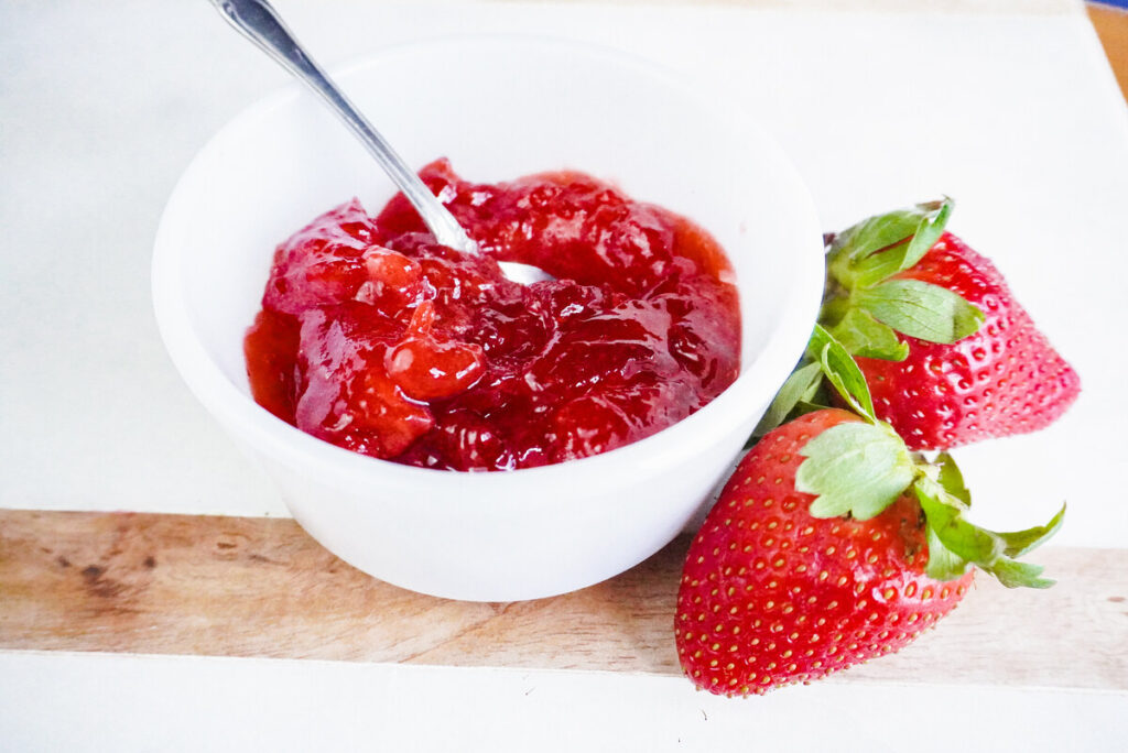 bowl of easy low sugar strawberry jam with 2 strawberries next to it on a marble background. 