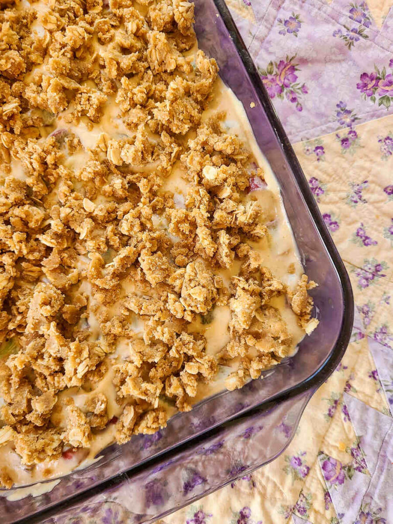 close up photo of unbakedRhubarb Crumble Cream Cheese Bars in purple pan set on a yellow and purple floral square quilt. 