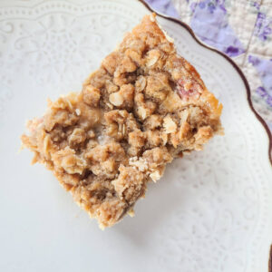 rhubarb crumble cream cheese bar square set on a scalloped white plate with a purple quilt background