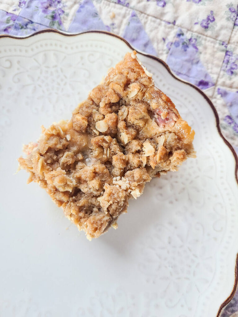 rhubarb crumble cream cheese bar square set on a scalloped white plate with a purple quilt background