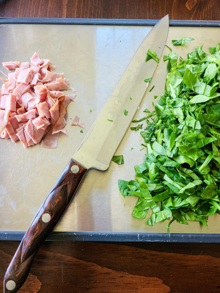 cutting board with sliced ham and spinach one it next to a large chef knife on a stainless steel cutting board. 
