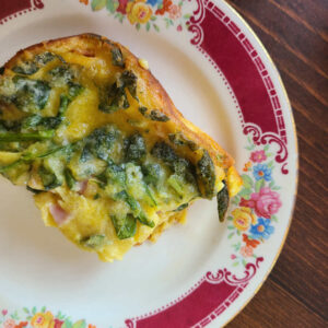 square slice of savory German pancake yellow with green pieces in it on a red floral plate with a dark wood background.