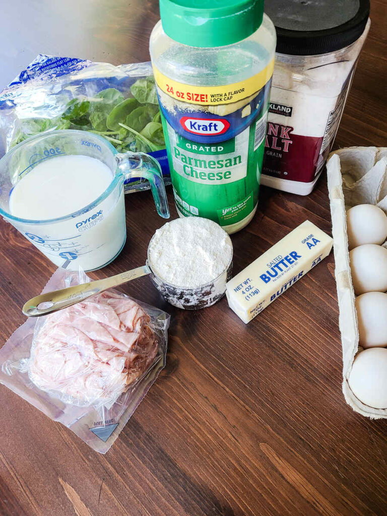 ingredients for savory dutch baby pancakes laid out of dark wood table. spinach, milk, parmesan cheese, pink salt, eggs, flour, butter, and ham lunchmeat 