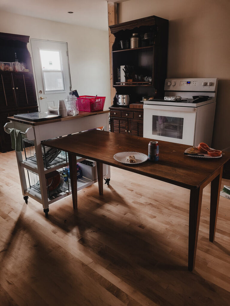 kitchen with white walls, dark bookshelf with microwave, toaster, glass storage, wood table in the center of the room, white stove, white kitchen cart with drawers. 