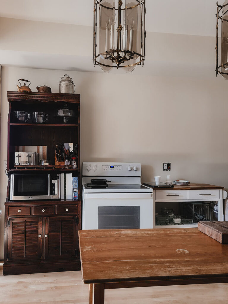 kitchen with white walls, dark bookshelf with microwave, toaster, glass storage, wood table in the center of the room, white stove, white kitchen cart with drawers. 