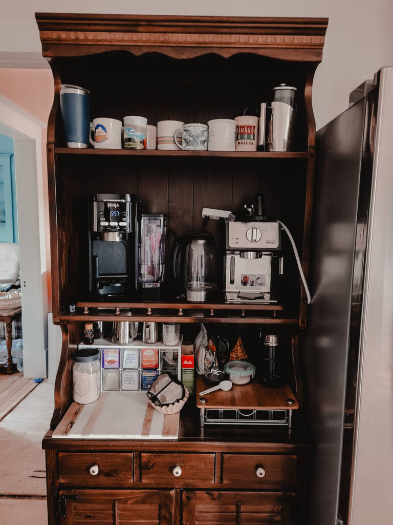 dark wood kitchen hutch with coffee mugs and makers, stainless steel fridge to the right of the hutch. white walls, light flooring 