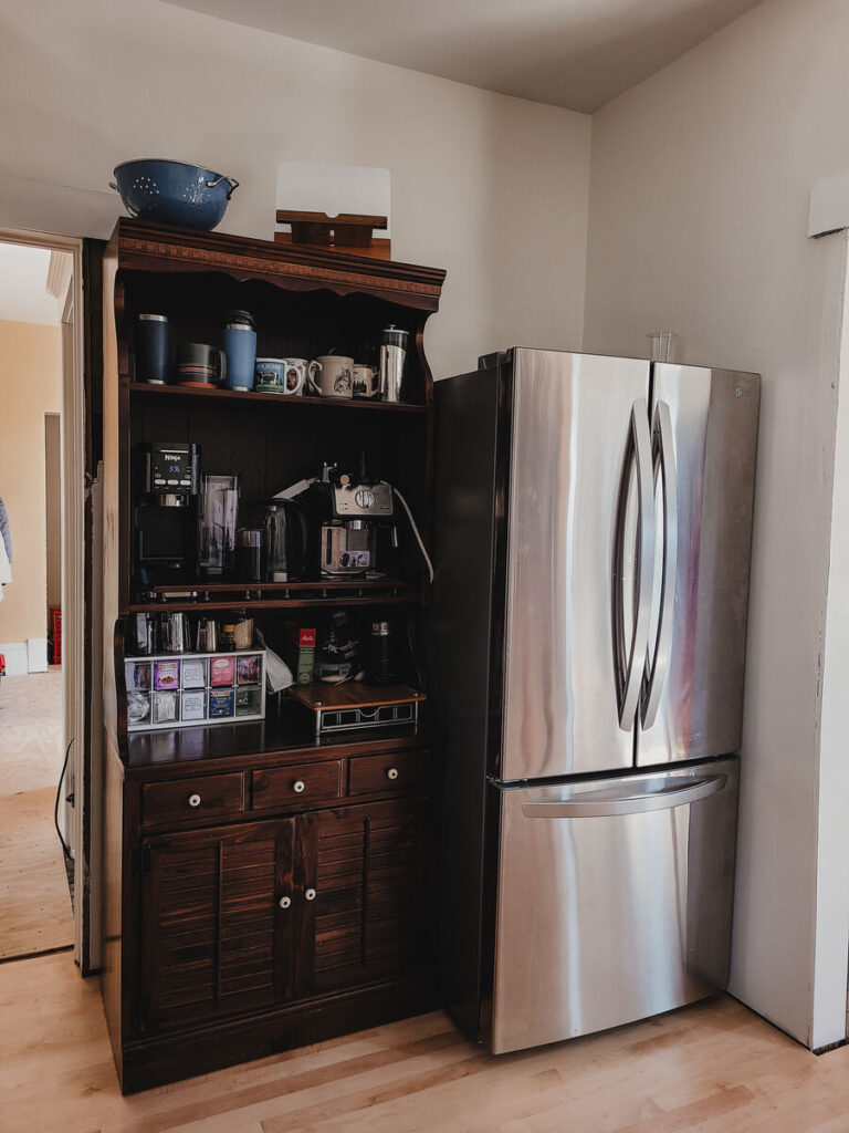 dark wood kitchen hutch with coffee mugs and makers, stainless steel fridge to the right of the hutch. white walls, light flooring 