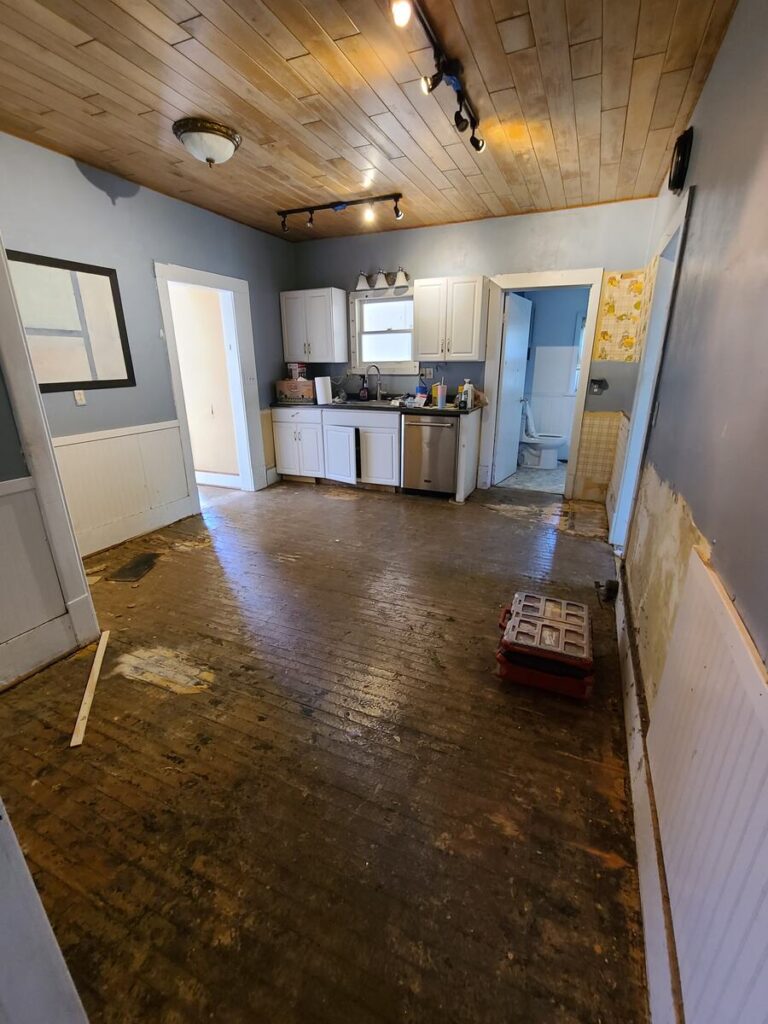 empty dirty kitchen with wood ceiling, blue walls, white trim and wall panels, and a dark wood floor.  counter with dishwasher and kitchen sink. 