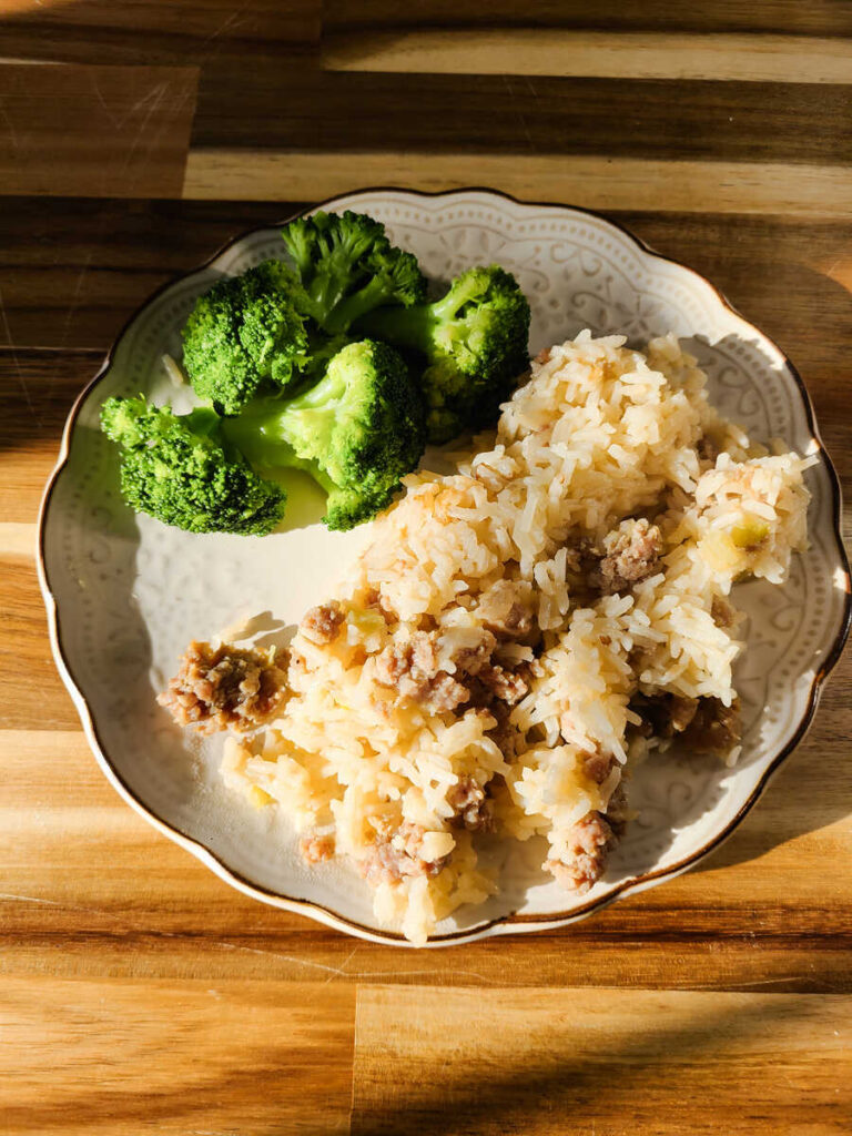 Simple Sausage Rice Dinner Instant Pot and Stovetop Recipe. wood block background with cream scalloped dinner plate full of sausage rice with broccoli on the side.