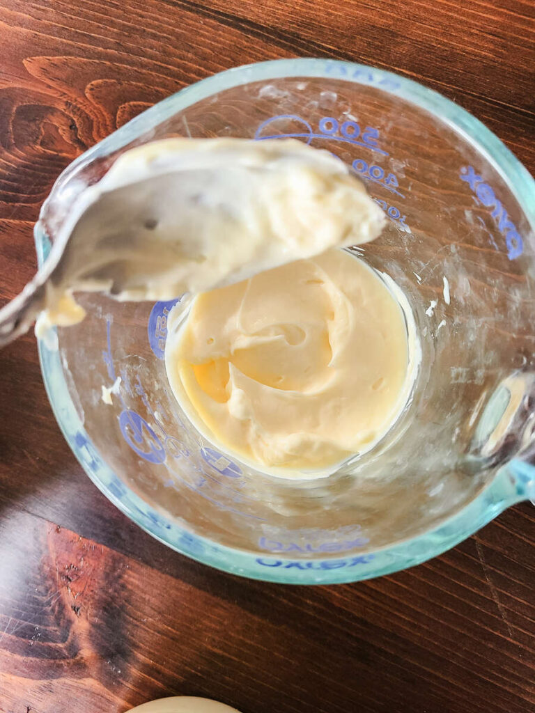 woman measuring mayo in liquid measuring cup using a metal spoon  to make coleslaw dressing. 