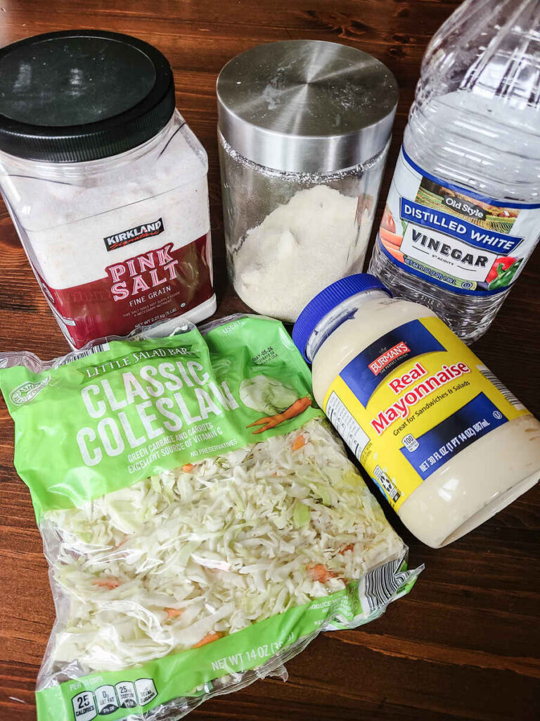 Ingredients for making the best easy coleslaw recipe coleslaw mix, pink salt, mayo, vinegar and sugar on wood table 