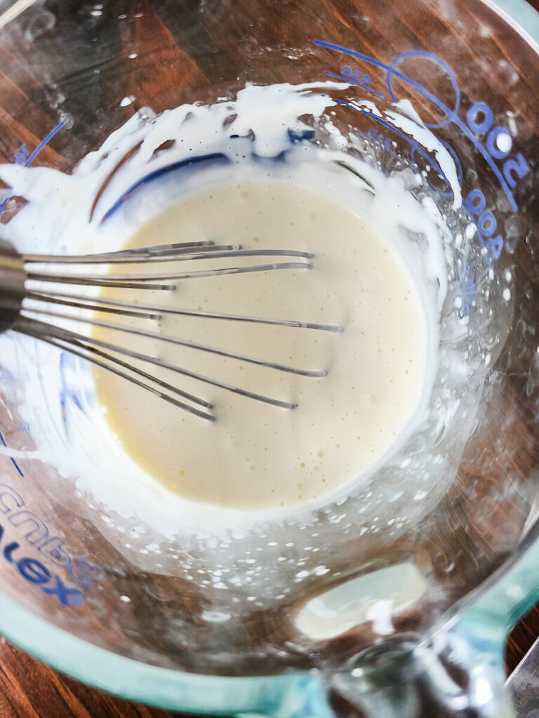 woman mixing white creamy coleslaw dressing in liquid measuring cup with metal whisk. 