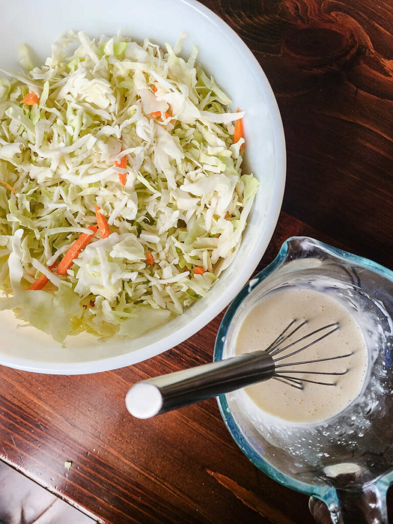 bowl of cabbage mix sitting next to cup of creamy coleslaw dressing in liquid measuring cup on dark wood table. 