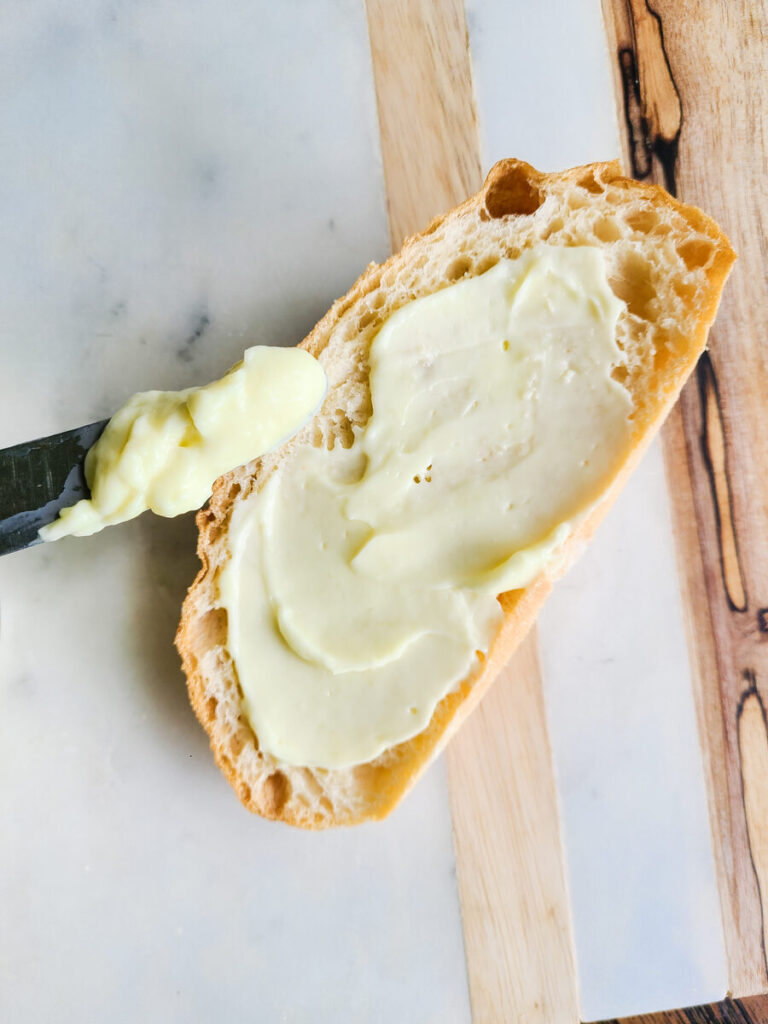 slice of French bread slathered with egg white mayo on a marble cutting board with a butter knife dipped into mayo. 