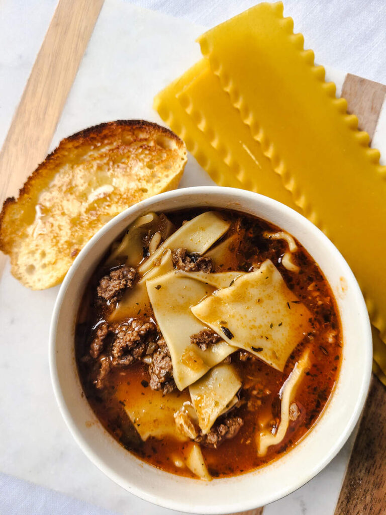 bowl with lasagna soup with French bread slice next to it and uncooked whole lasagna noodles all on marble cutting board background 