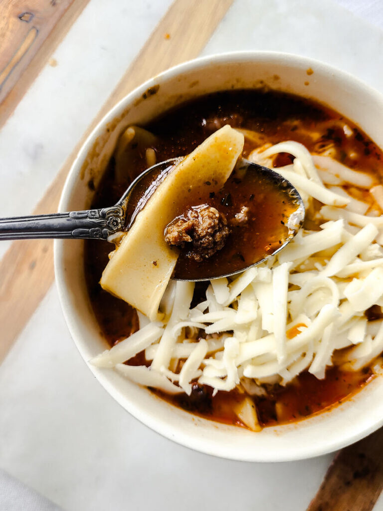 bowl of hot lasagna soup with cheese on top. with a spoon dipped into it on a marble tray. 