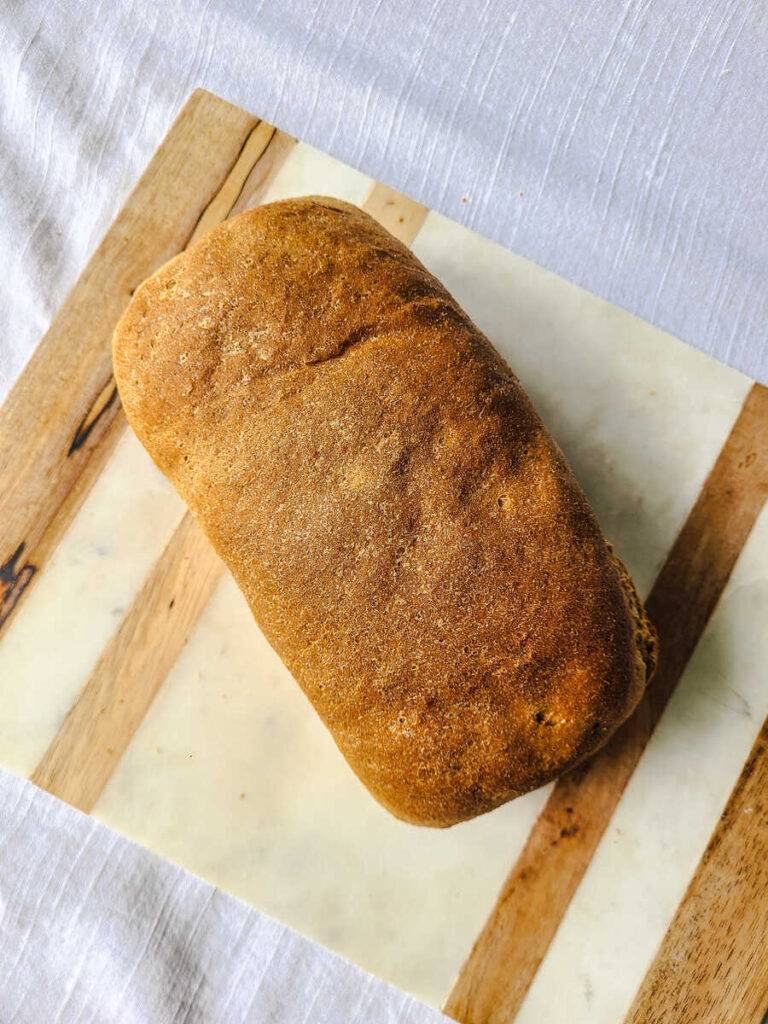 whole loaf of whole wheat bread sitting horizontally on marble and wood cutting board 