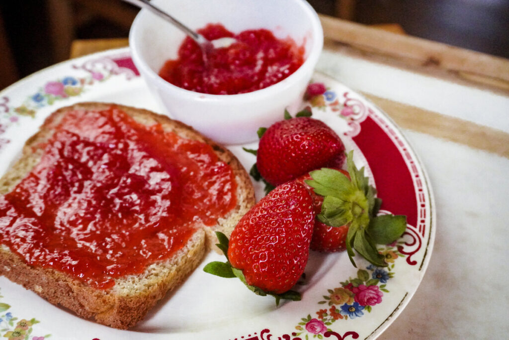 slice of whole wheat bread slathered in strawberry jam with strawberries and white bowl of strawberry jam sitting next to the slice of bread. 