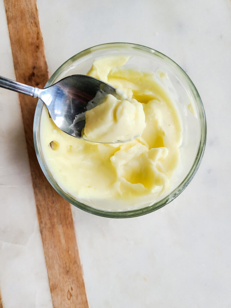 marble and wood backround with small glass bowl of homemade easy egg white mayo in it with a silver spoon dipped into it