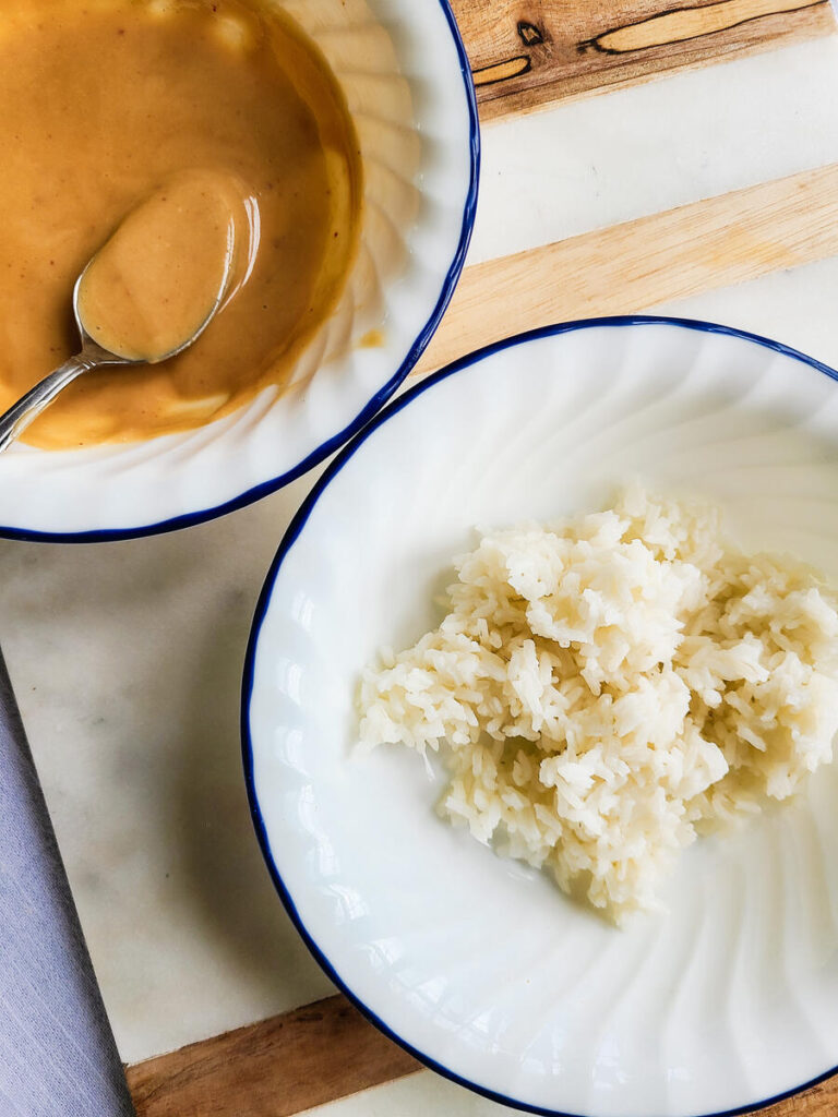 background of a marble and wood counter top with 2 white bowls with dark blue edge sitting next to each other one full of spicy kimchi mayo with a spoon and one full of cooked white rice.